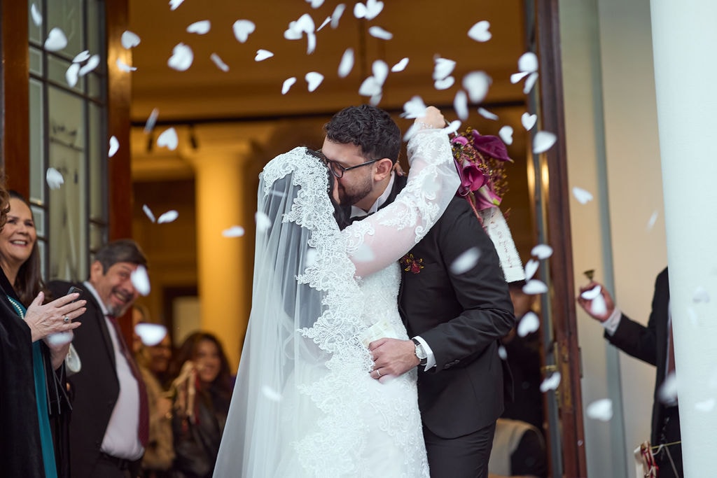 Los novios besándose a la salida de la iglesia bajo una lluvia de pétalos, con los invitados alrededor