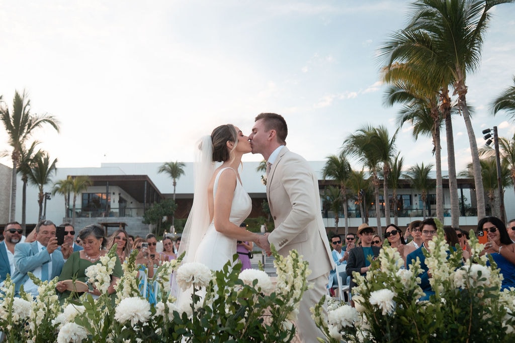 Los novios besándose en la playa bajo las palmeras, con los invitados alrededor