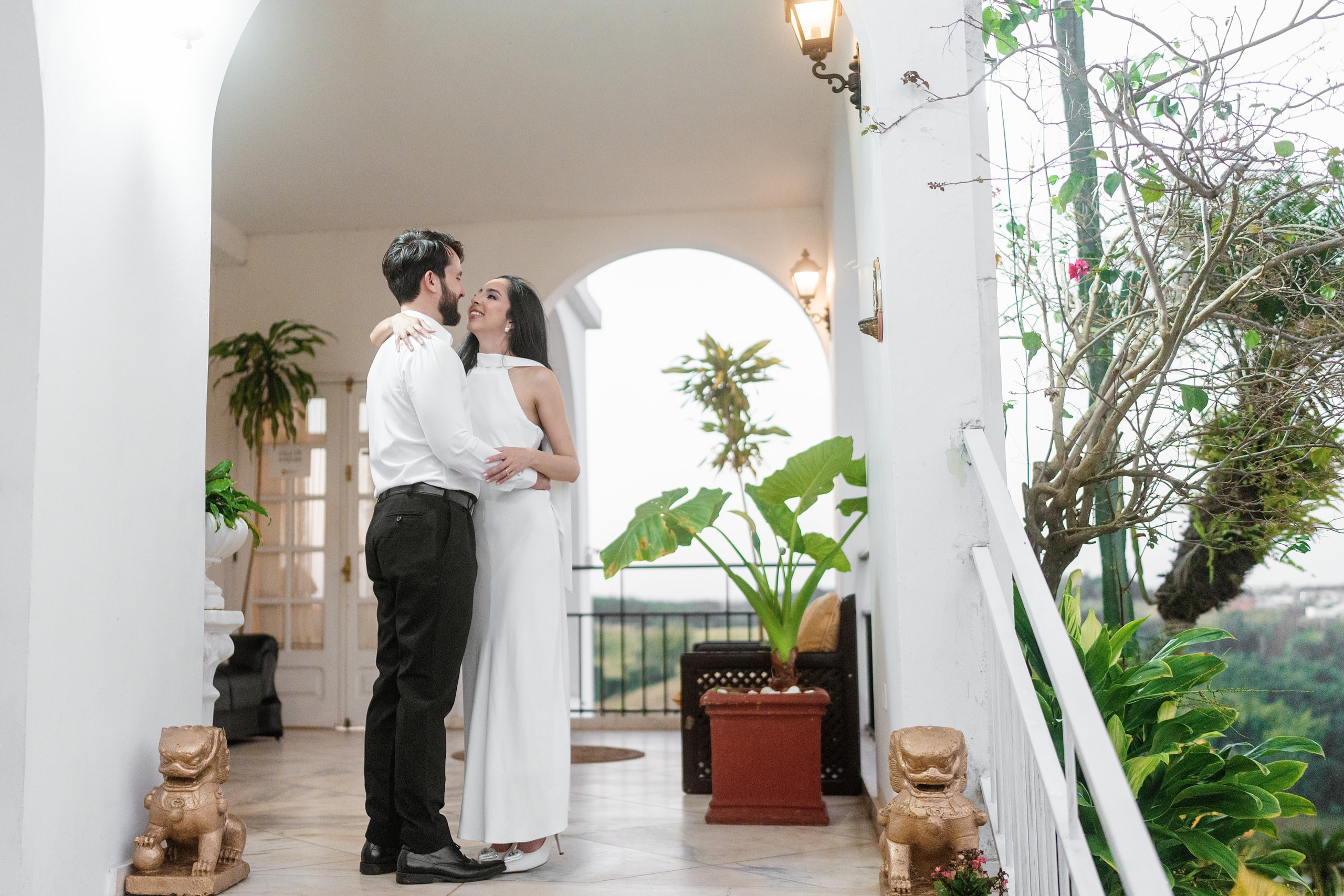 La pareja abrazada en una galería de arcos blancos con plantas, ella de vestido blanco largo