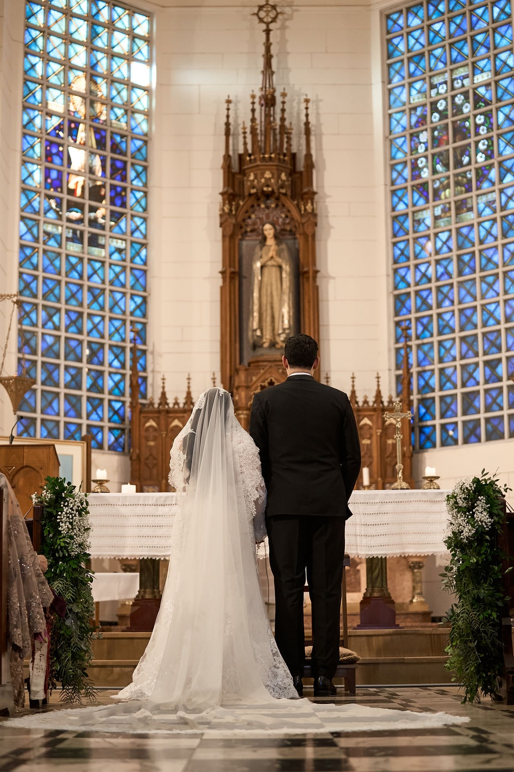 Los novios de espaldas ante el altar de la iglesia, el velo largo cayendo hasta el piso ajedrezado, entre los vitrales azules