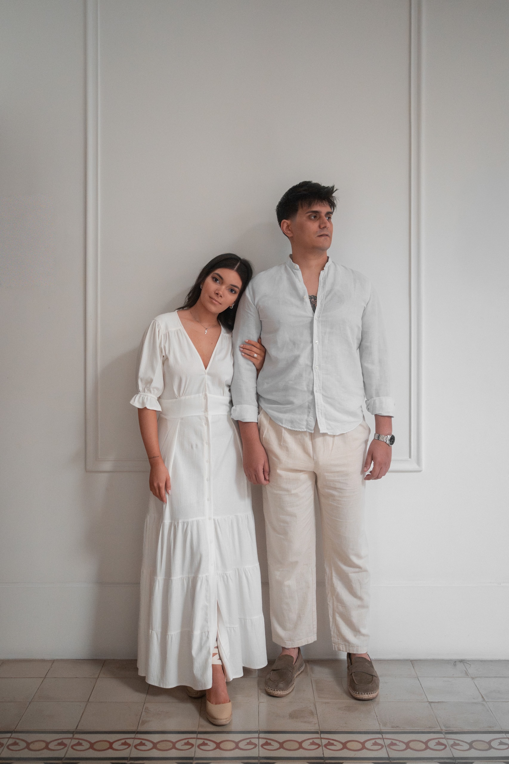 The couple standing against a white wall: she in a long white dress, he in a light linen shirt and beige trousers