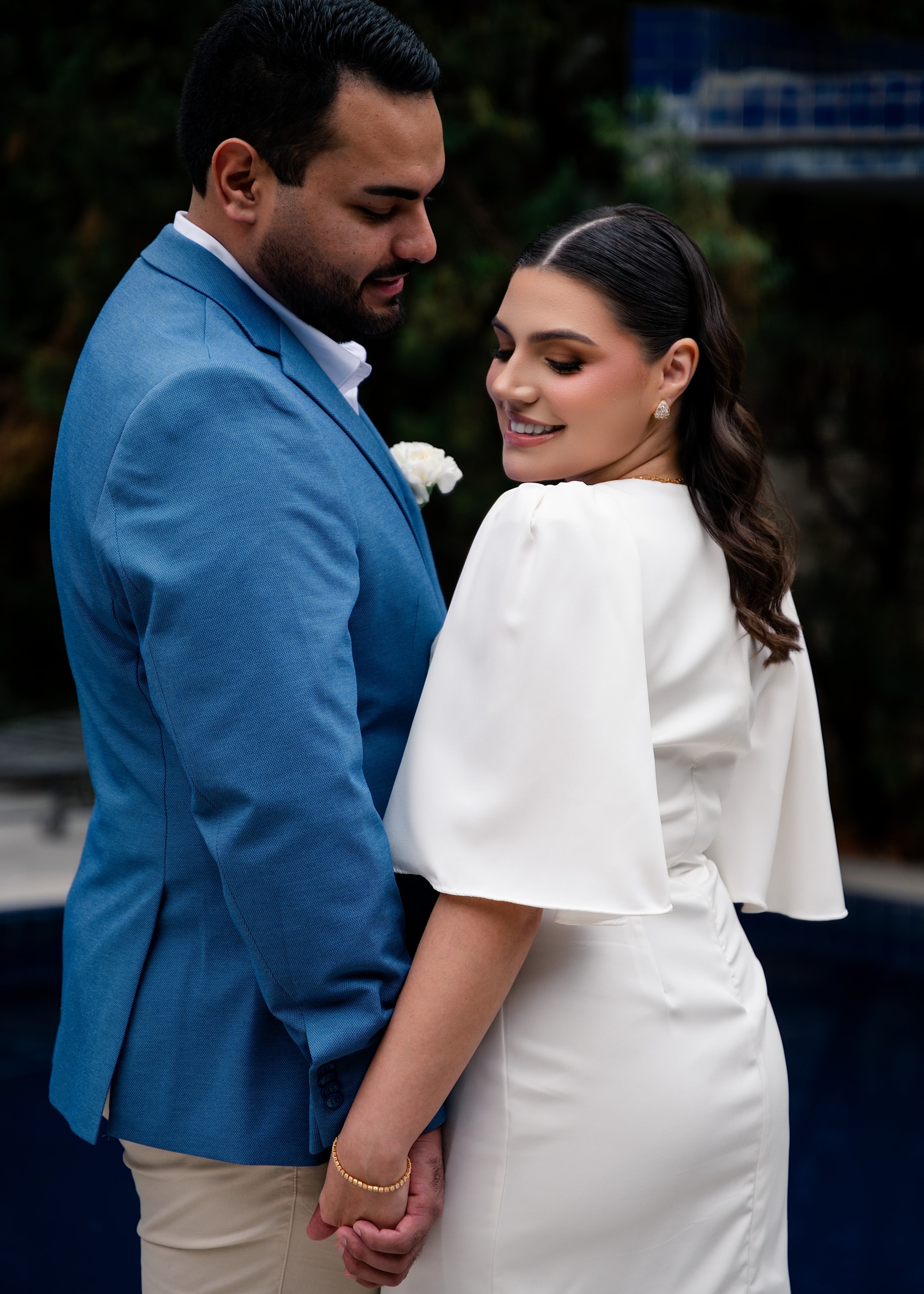 A couple holding hands; he in a light-blue blazer, she in a white dress looking over her shoulder