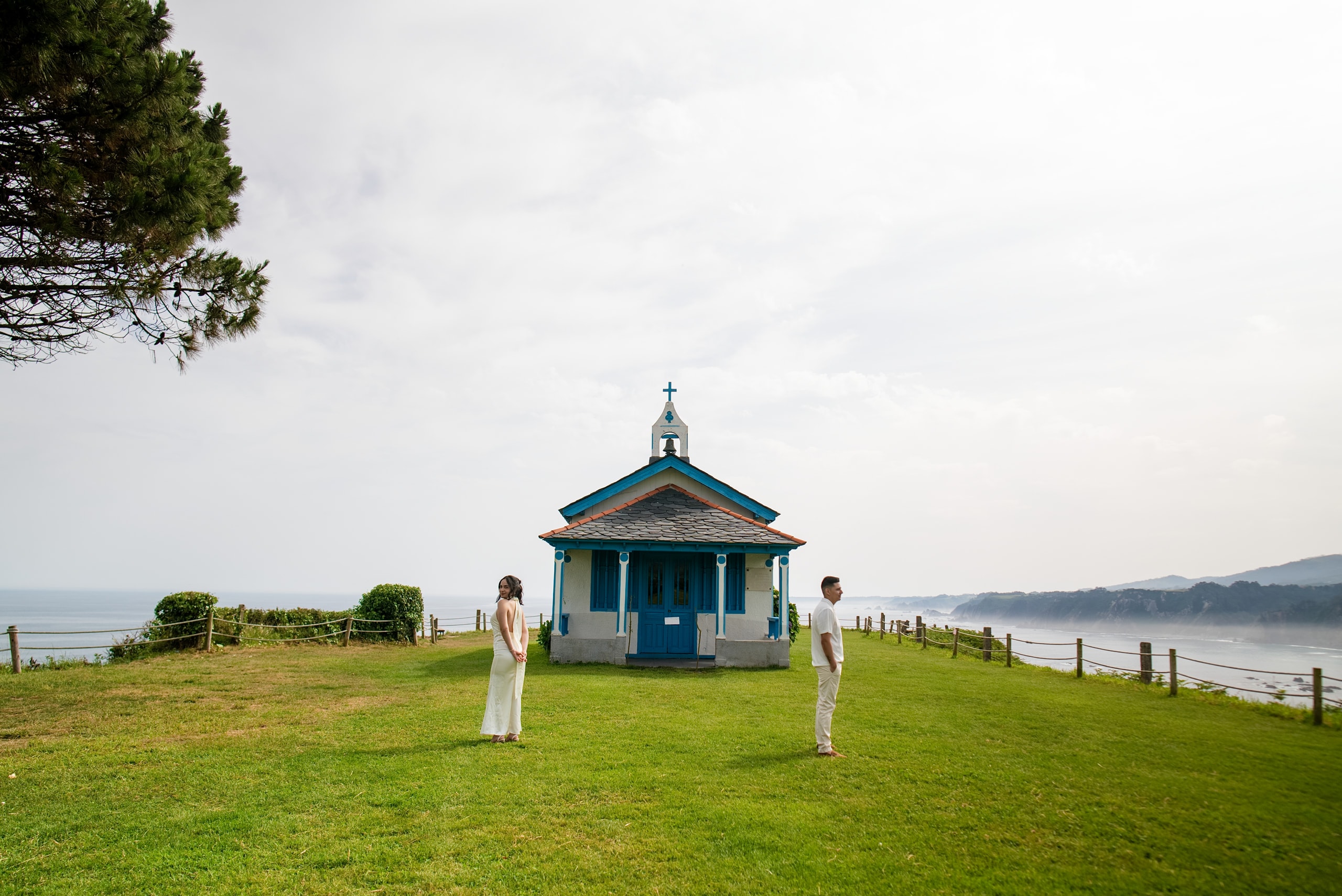Los novios de pie en el pasto frente a una capilla azul y blanca, con el mar detrás