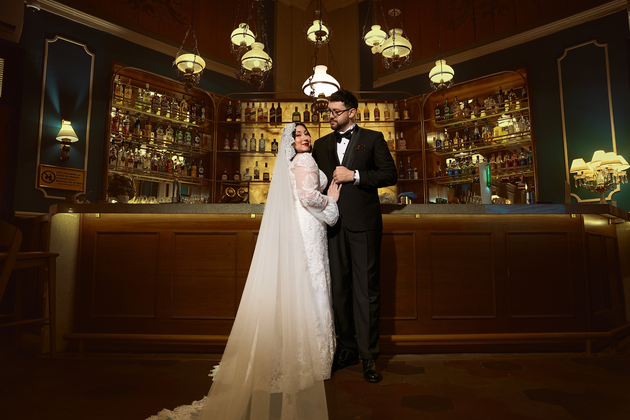 The newlyweds before the lit bar of a classic saloon, she in lace and veil, he in a tuxedo