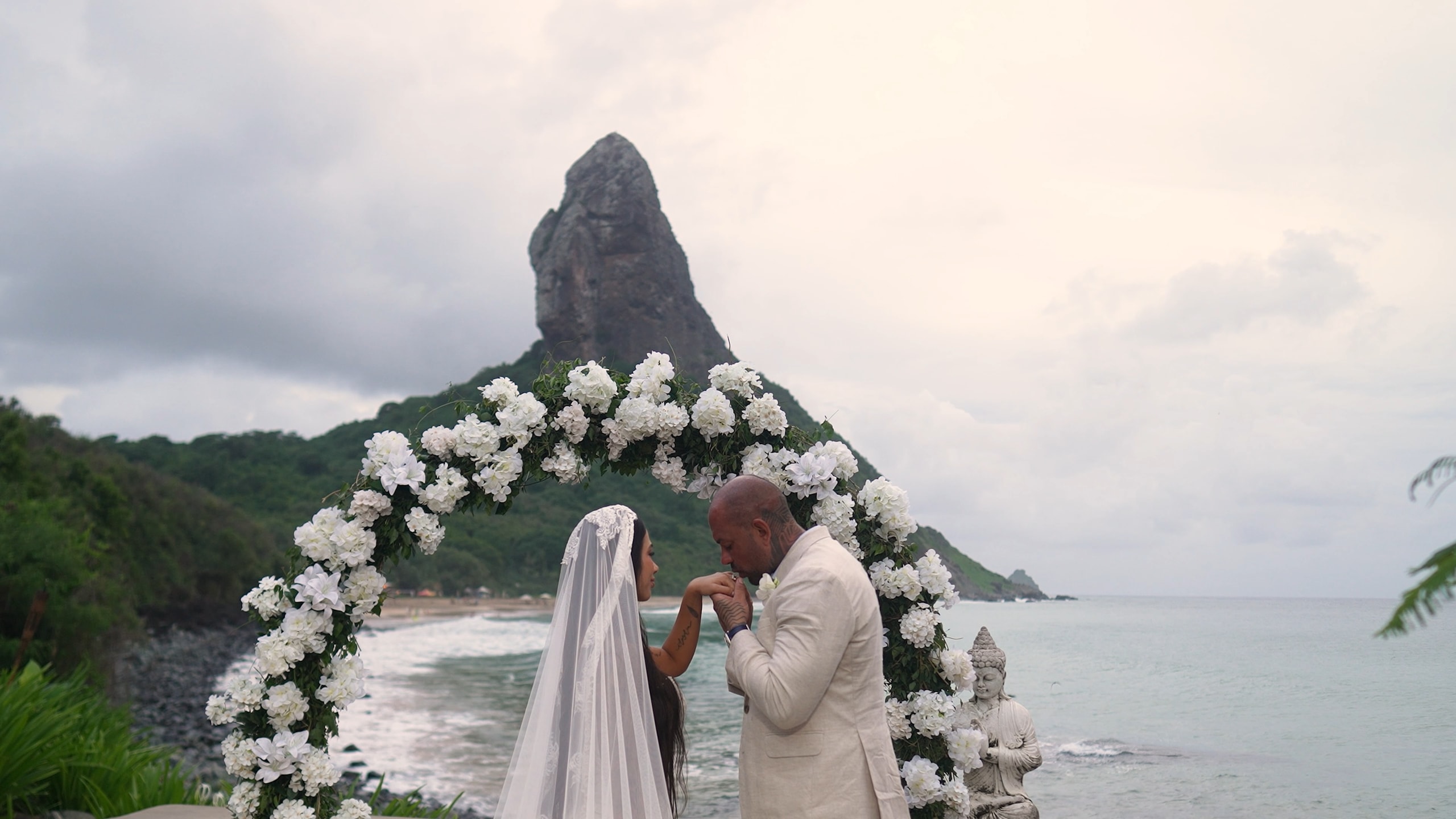 Los novios bajo un arco de flores blancas frente al mar, con el Morro do Pico de Fernando de Noronha detrás