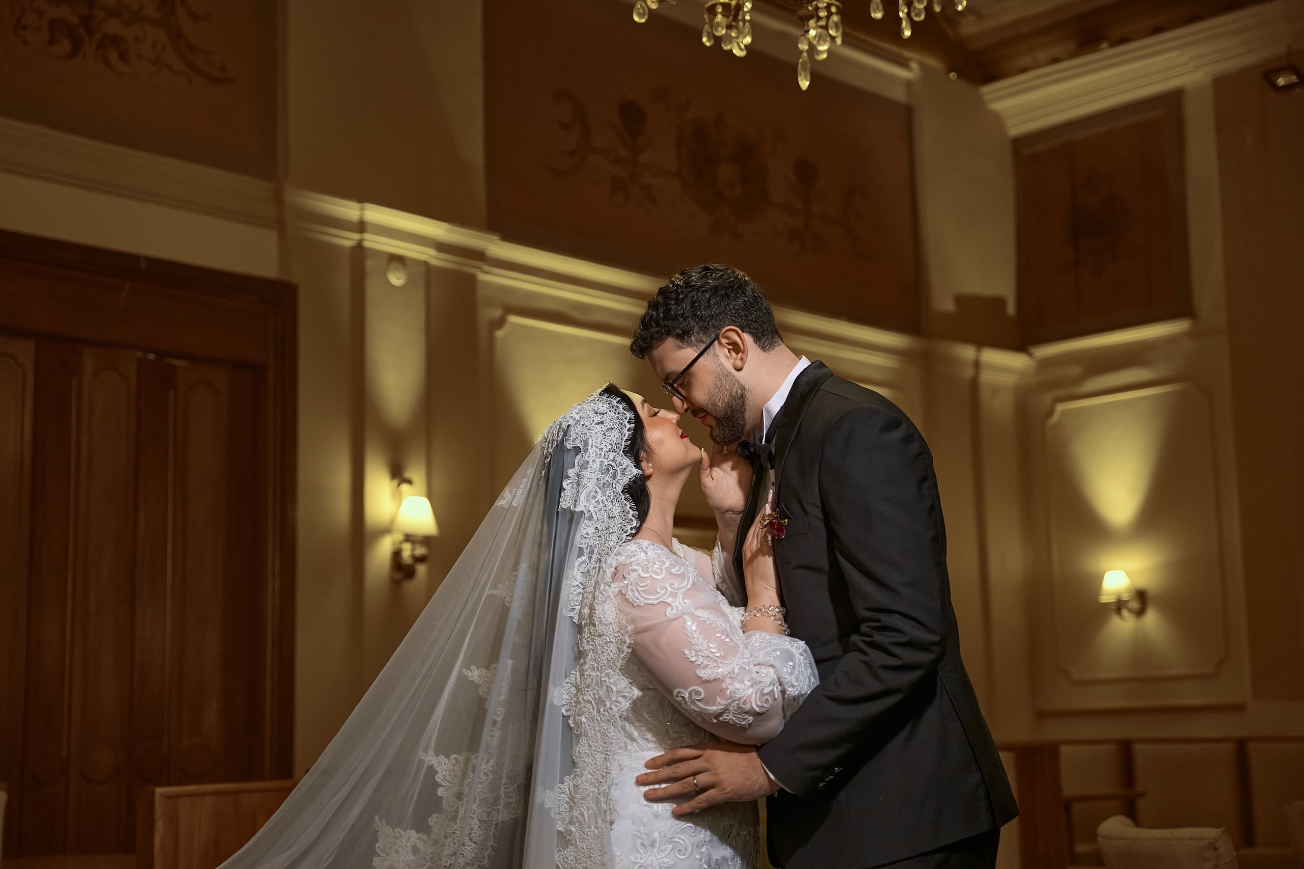 The couple about to kiss in a room of gilded moldings, she in a lace mantilla and he in a tuxedo