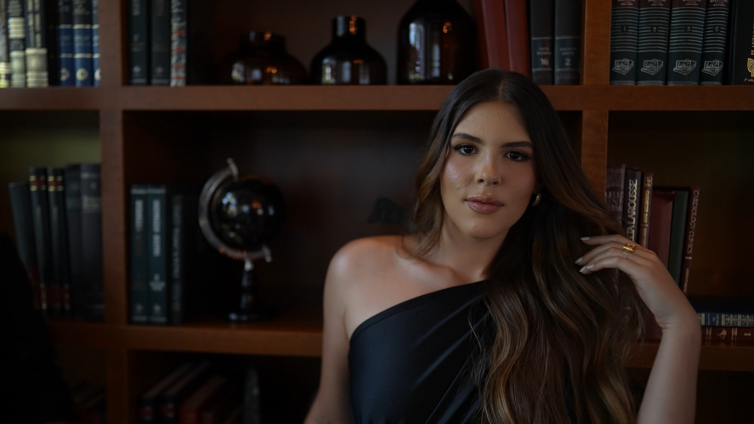 The bride seated in the hotel library, in a black one-shoulder dress, among wooden shelves and a globe