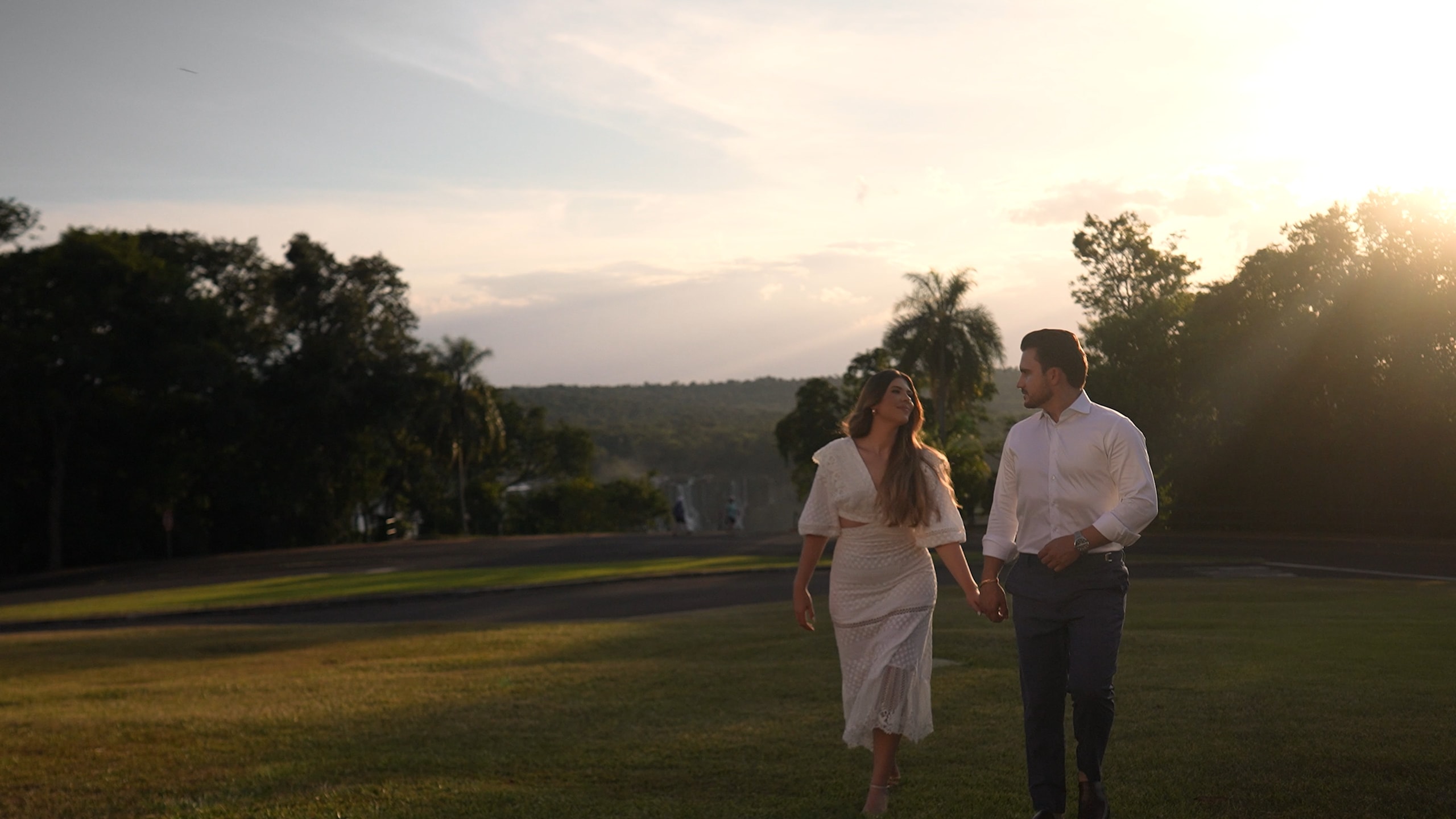 The couple walking hand in hand across the hotel's open lawn at sunset, among palm trees, golden light behind them