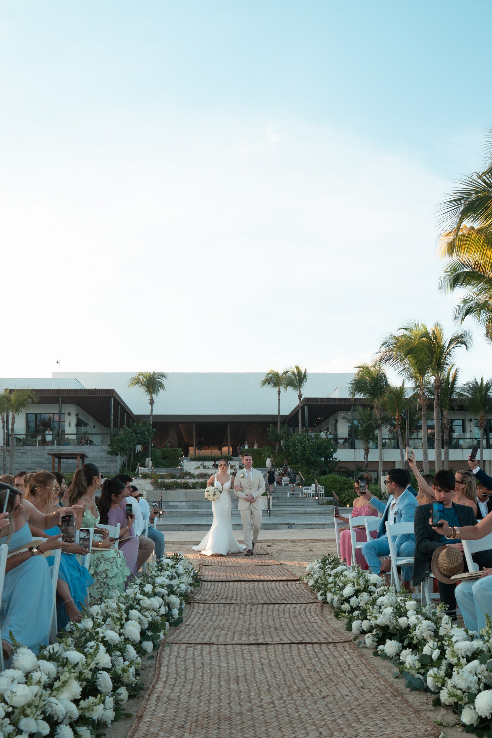 The couple walking down the ceremony aisle between palm trees and white flowers, facing the sea