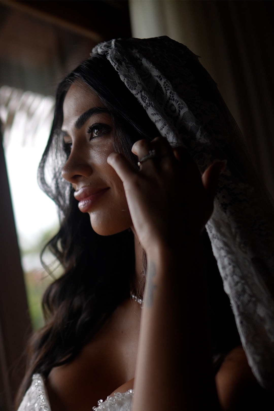 La novia de perfil con una mantilla de encaje sobre el pelo, tocándose la mejilla, a contraluz de una ventana
