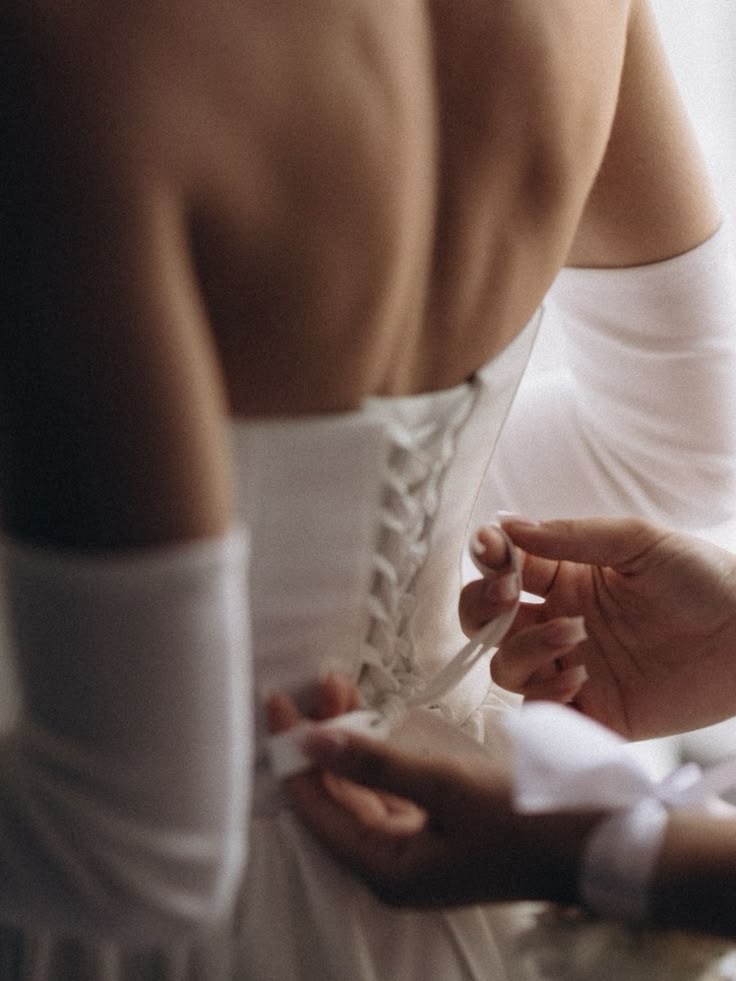Las manos de alguien atando el corsé del vestido en la espalda de la novia, con luz suave de ventana
