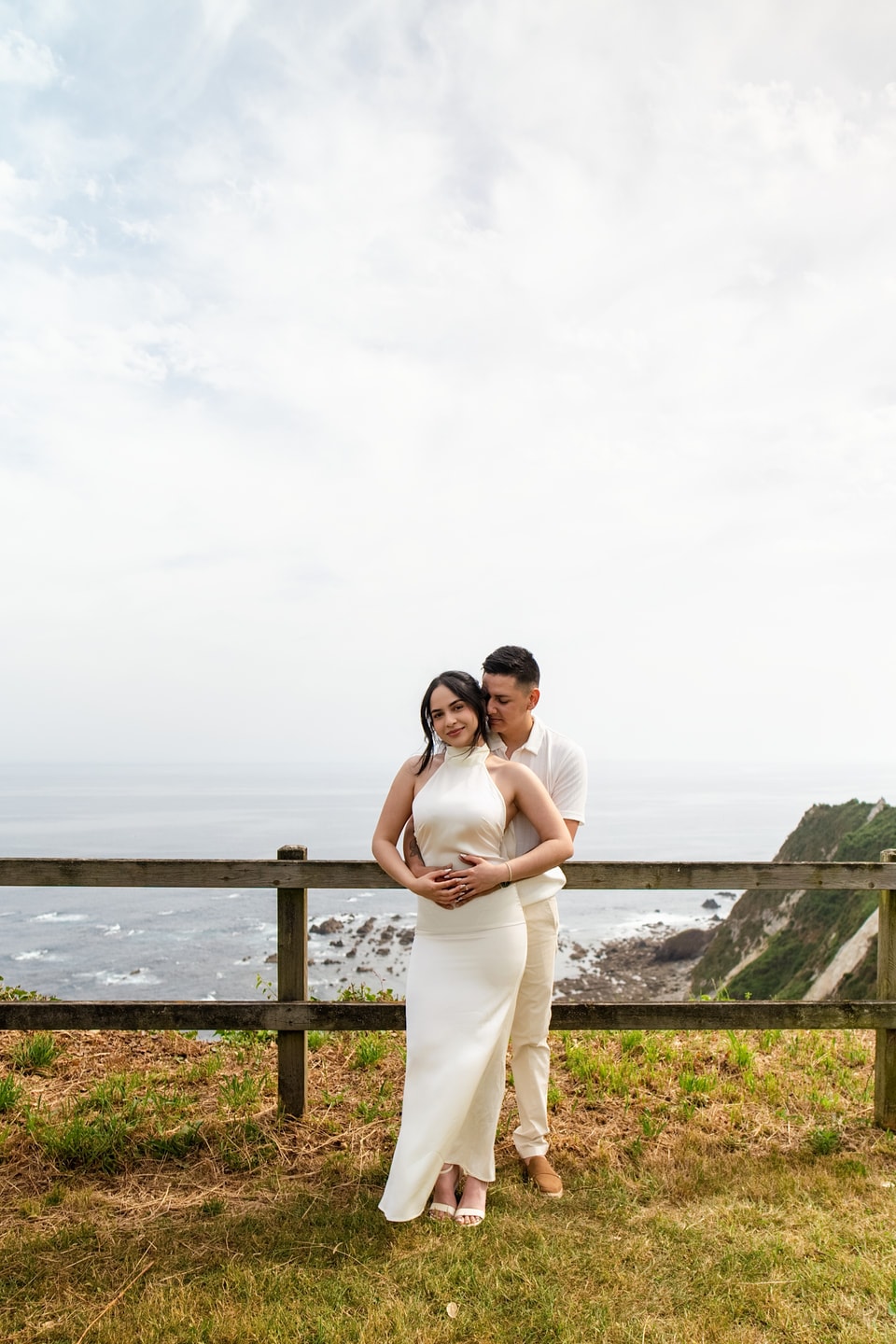 Los novios abrazados en un mirador sobre el mar de Asturias, los dos de blanco, con los acantilados verdes detrás
