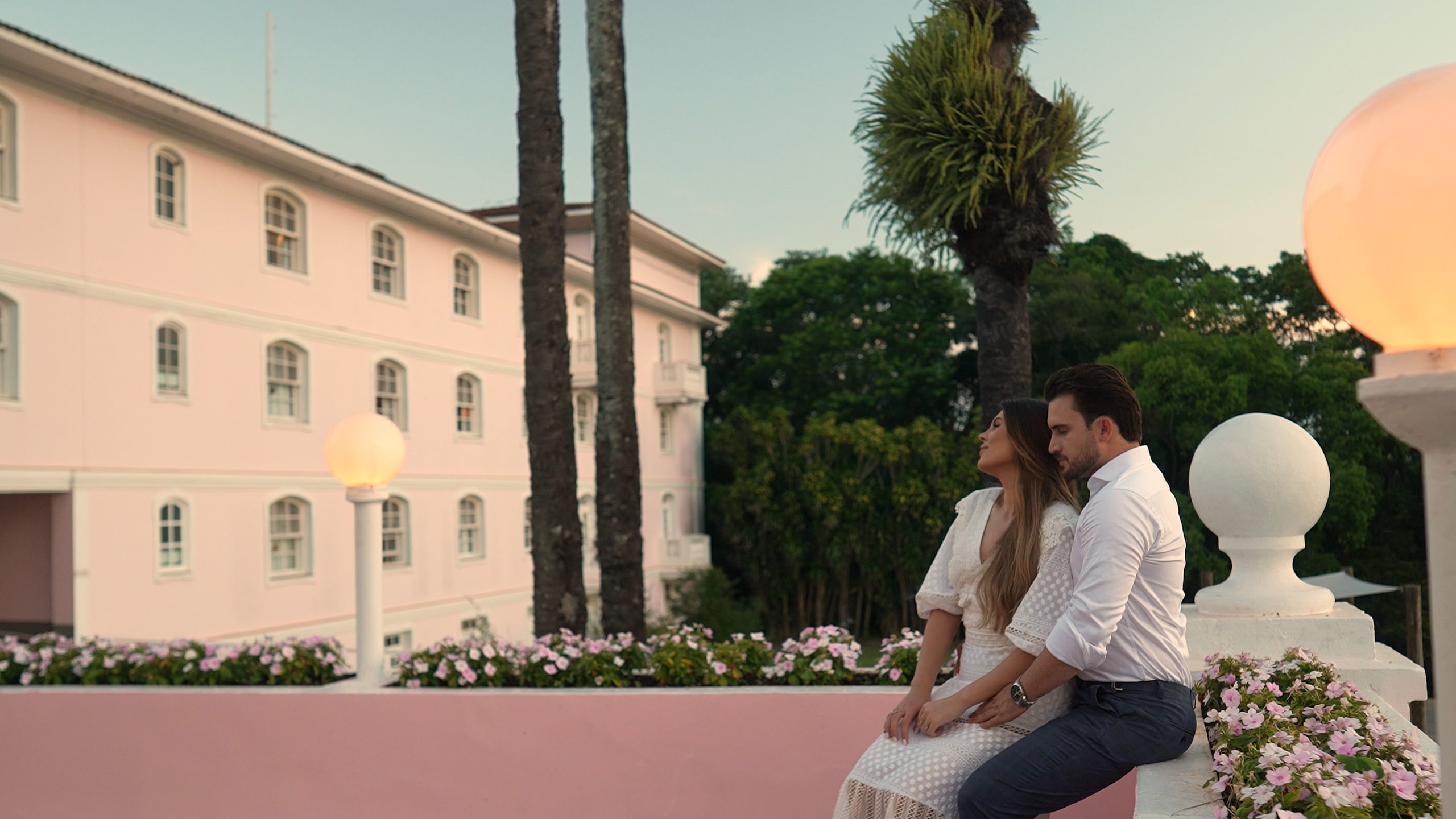 La pareja sentada en una balaustrada con la fachada rosa del hotel Belmond detrás, entre palmeras y flores