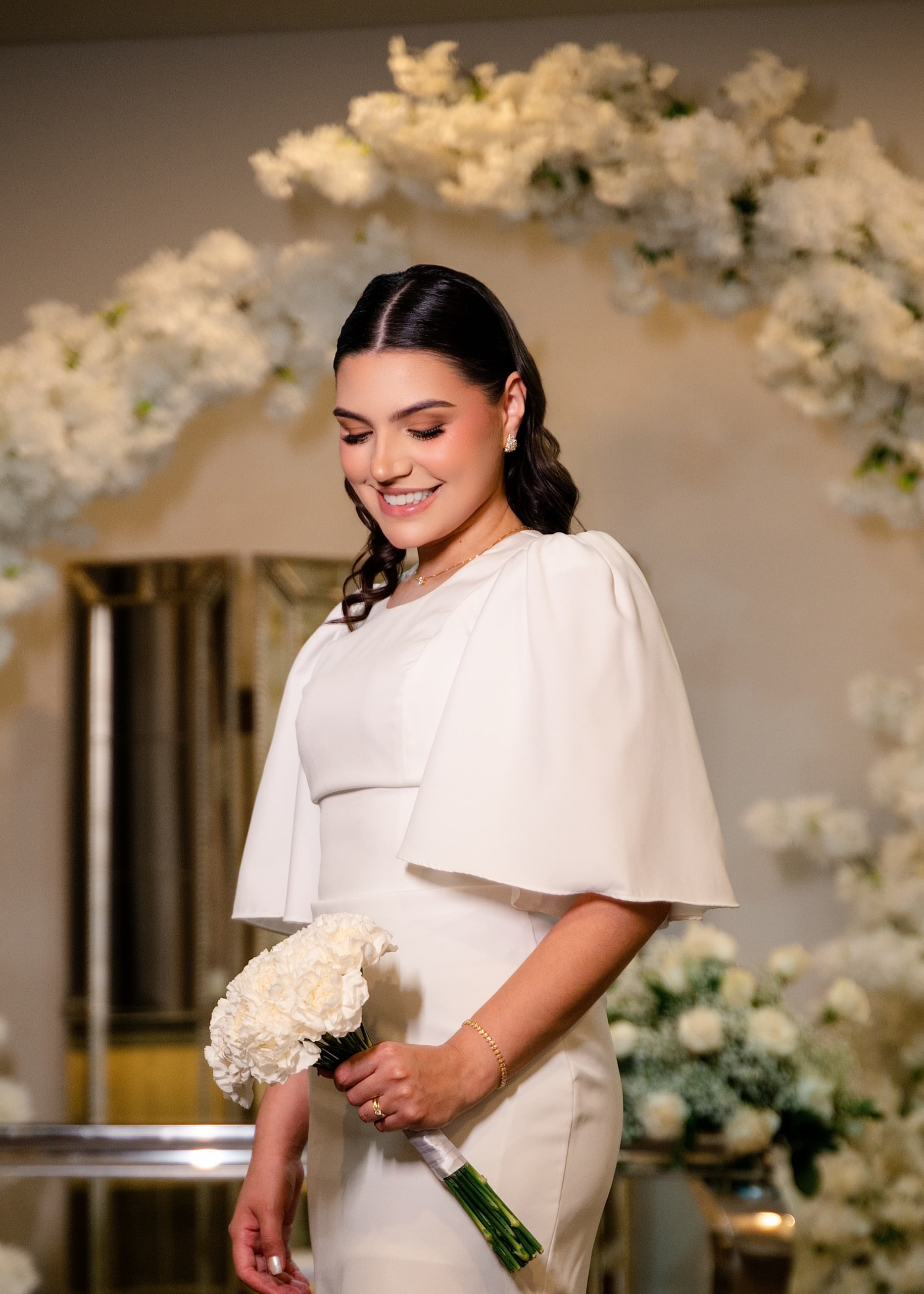 La novia de vestido blanco frente a un arco de flores blancas, con el ramo de claveles en las manos