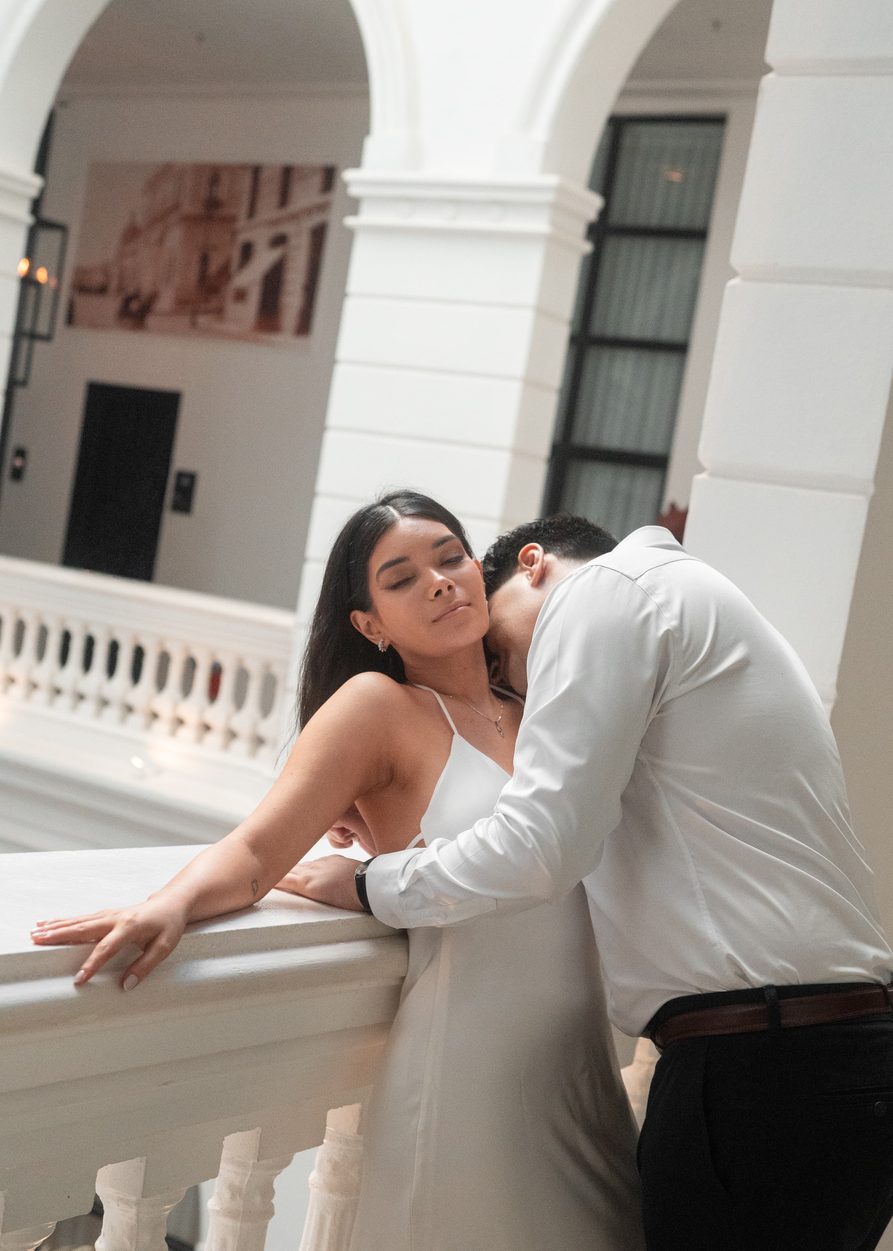 Los novios en la galería del hotel: ella apoyada de espaldas en la balaustrada y él inclinado hacia ella, entre columnas y arcos blancos