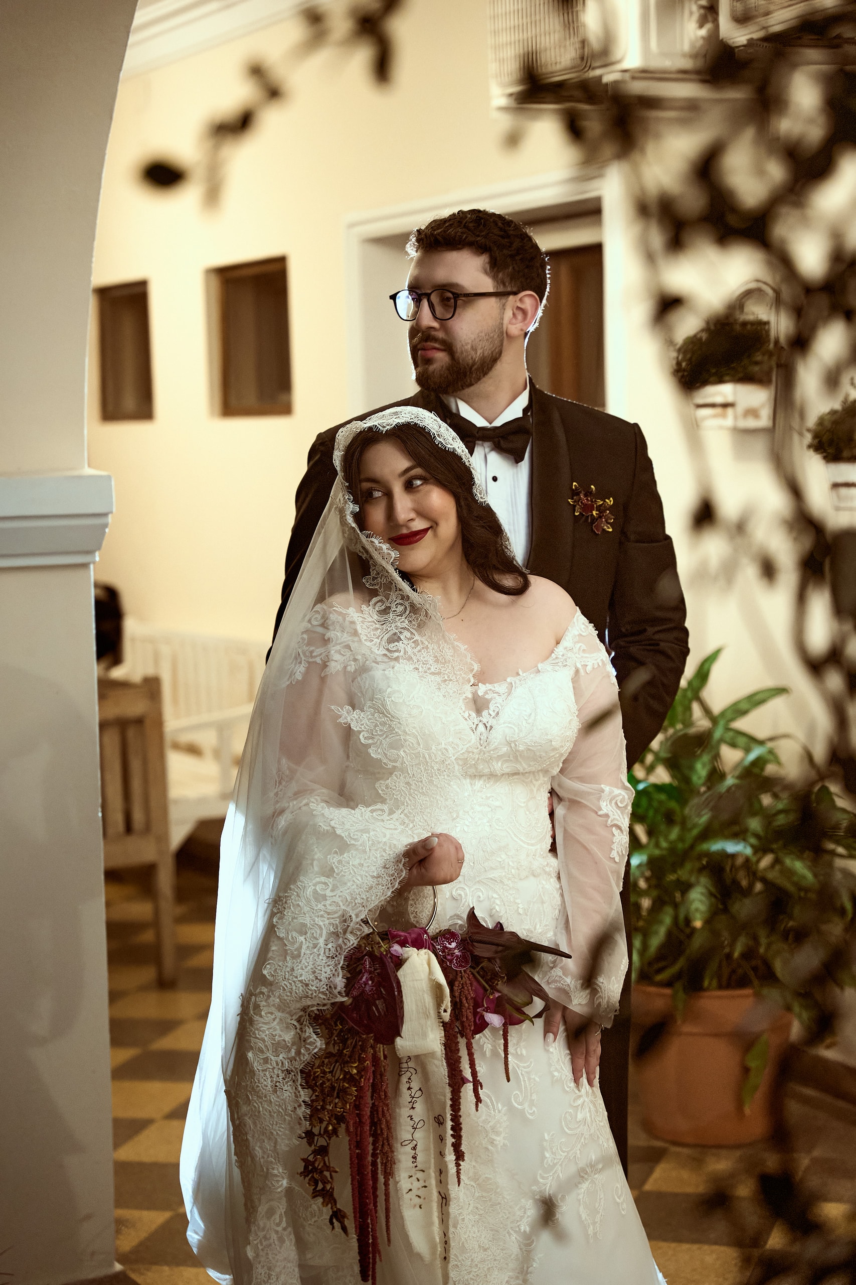 La novia con mantilla de encaje y un ramo de amaranto rojo, el novio de esmoquin detrás, en una galería de piso ajedrezado