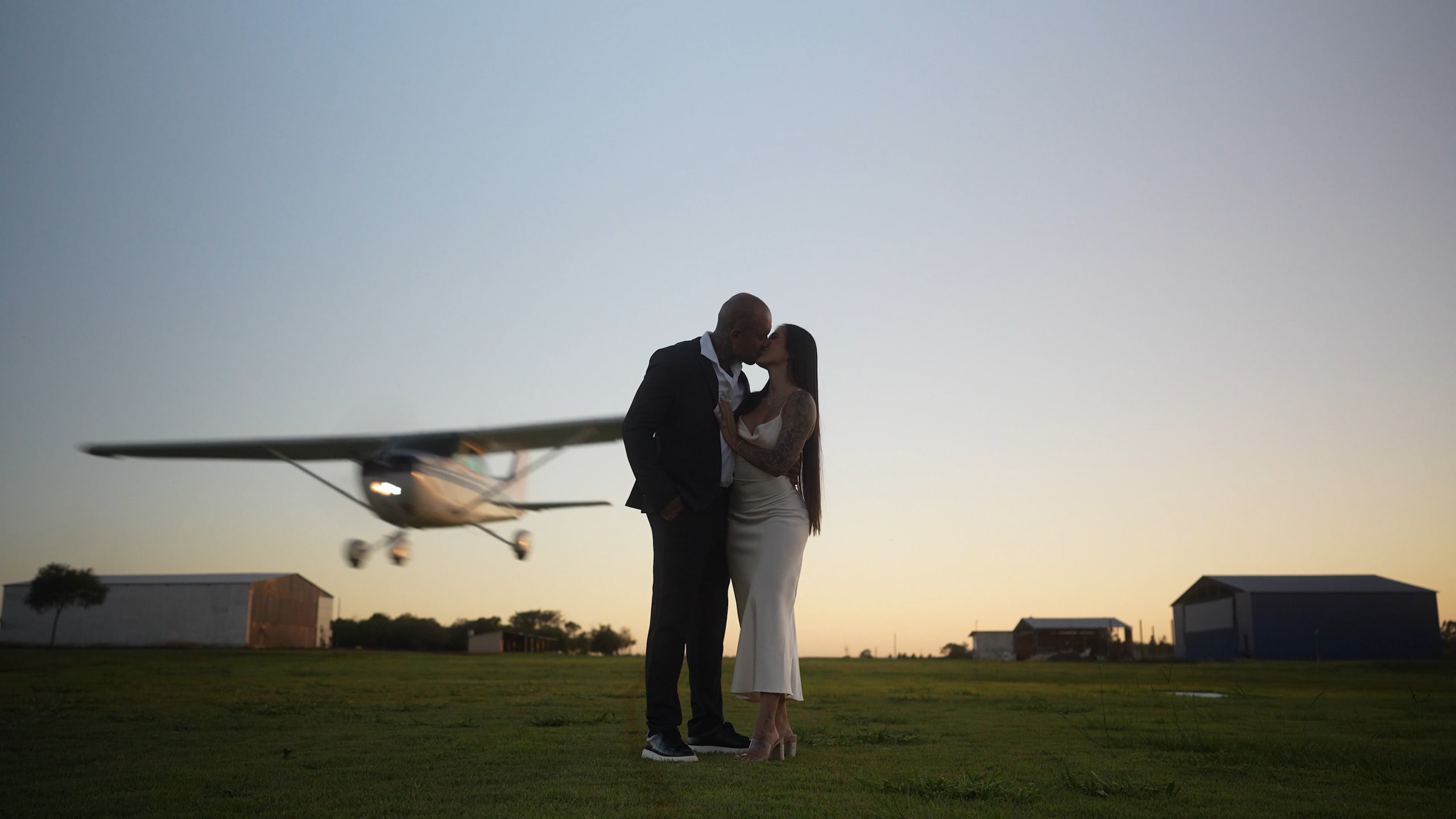 Una pareja besándose a contraluz junto a su avión, recortada contra el cielo dorado del atardecer