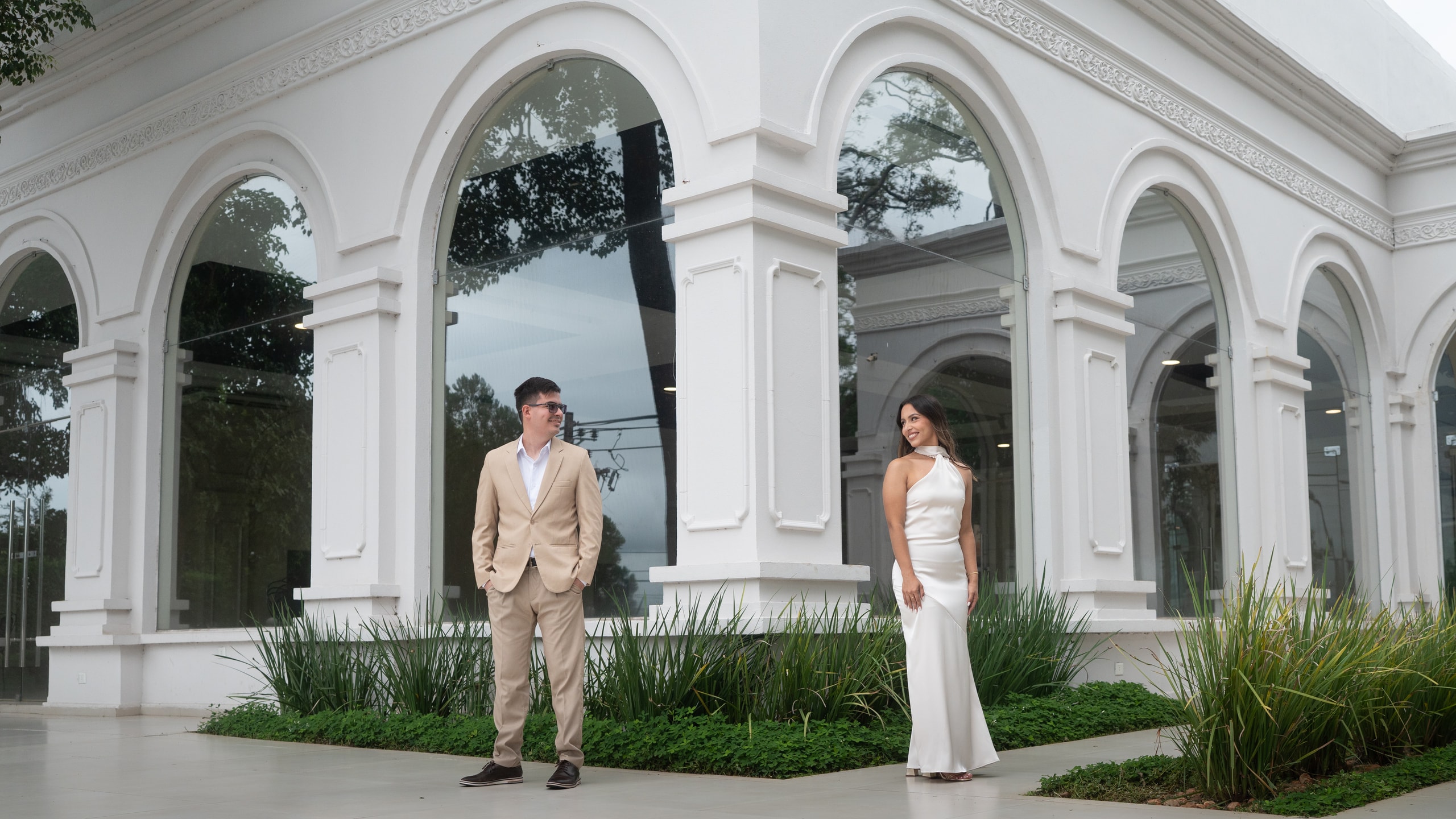 Una pareja en su pre-boda, separados bajo los arcos blancos de una galería: ella de vestido largo de satén claro, él de traje beige