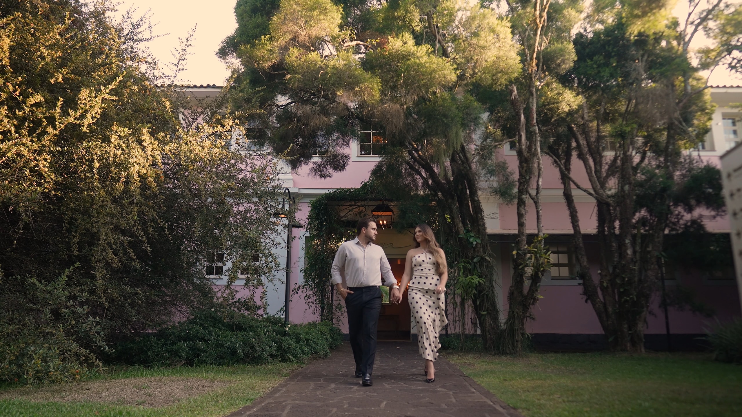 The couple walking hand in hand, both in white, with the hotel's pink facade and the garden behind them